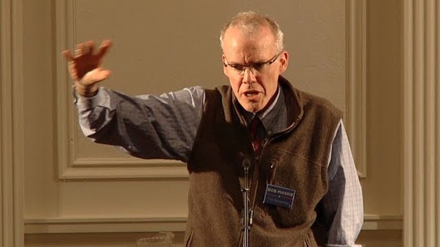 Bill McKibben, a light-skinned man wearing a brown vest, gives a talk at a podium.