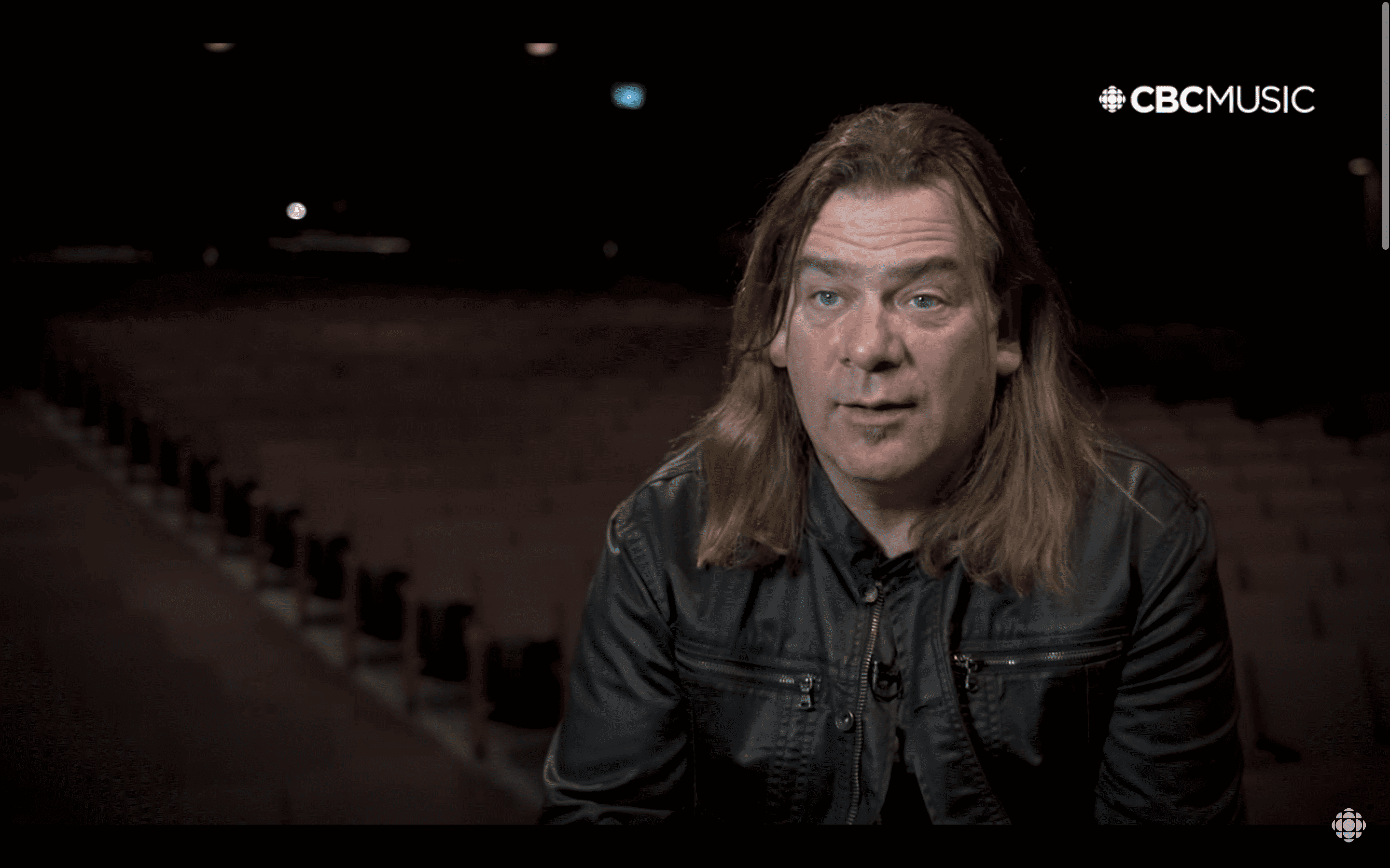 Alan Doyle, a light-skinned man with brown hair, giving an interview in an empty auditorium.